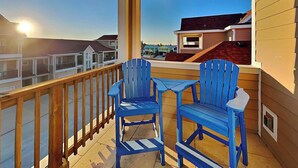 A balcony with two blue Adirondack chairs and a small table, overlooking a residential area with houses and a clear sky. - Just Beachy by Silver Sands Vacation Rentals (Port Aransas)