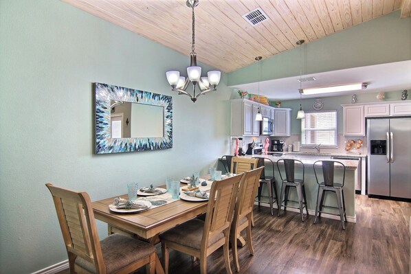 A dining area with a wooden table, chairs, and a chandelier. There is a kitchen area with cabinets and a refrigerator.