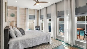 A bedroom with a bed, a ceiling fan, large windows with curtains, and a view of an outdoor pool. - Nauti Beach by Silver Sands Vacation Rentals (Port Aransas)