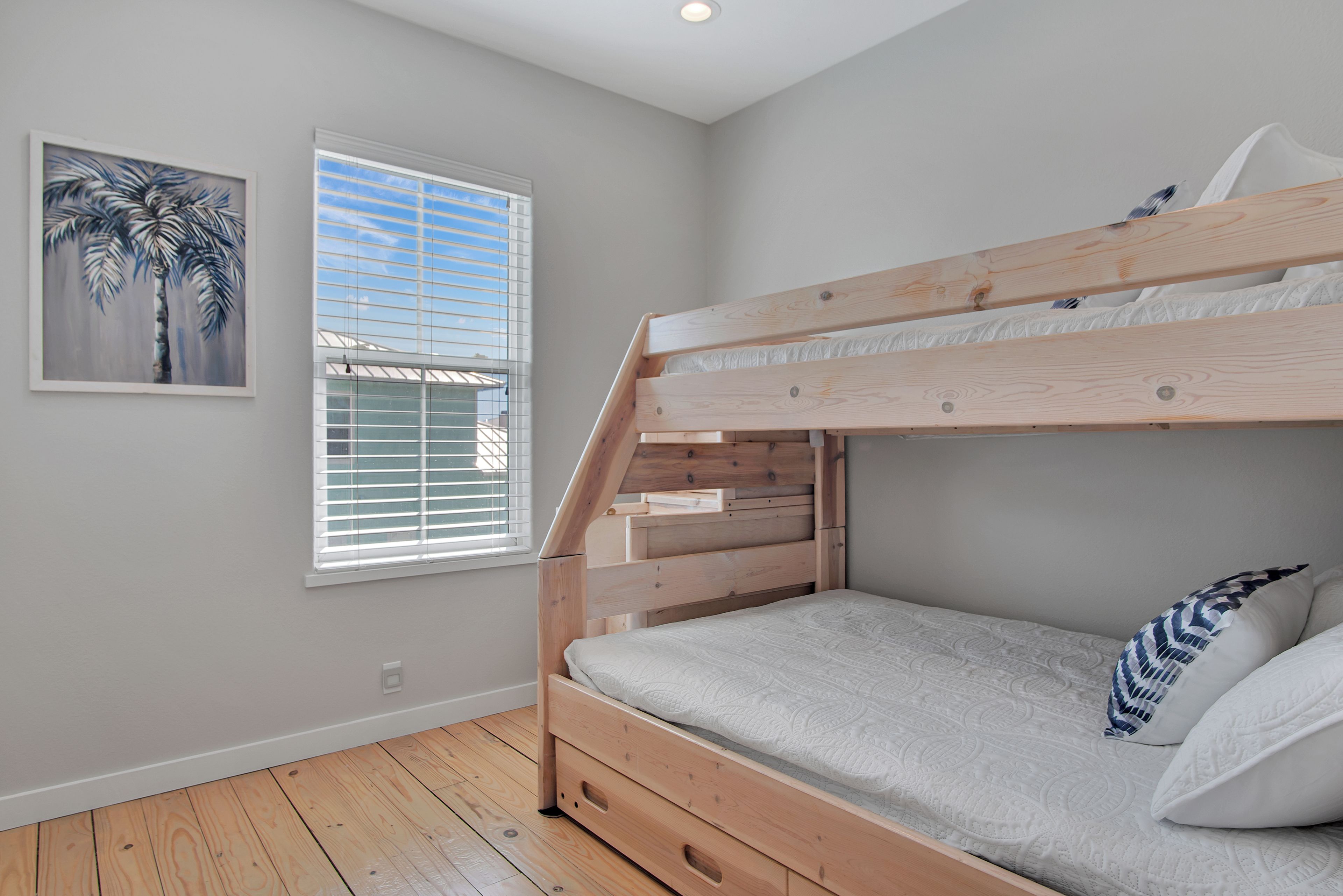 A bunk bed with a wooden frame, a window with blinds, and a painting of a palm tree on the wall.
