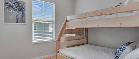 A bunk bed with a wooden frame, a window with blinds, and a painting of a palm tree on the wall.