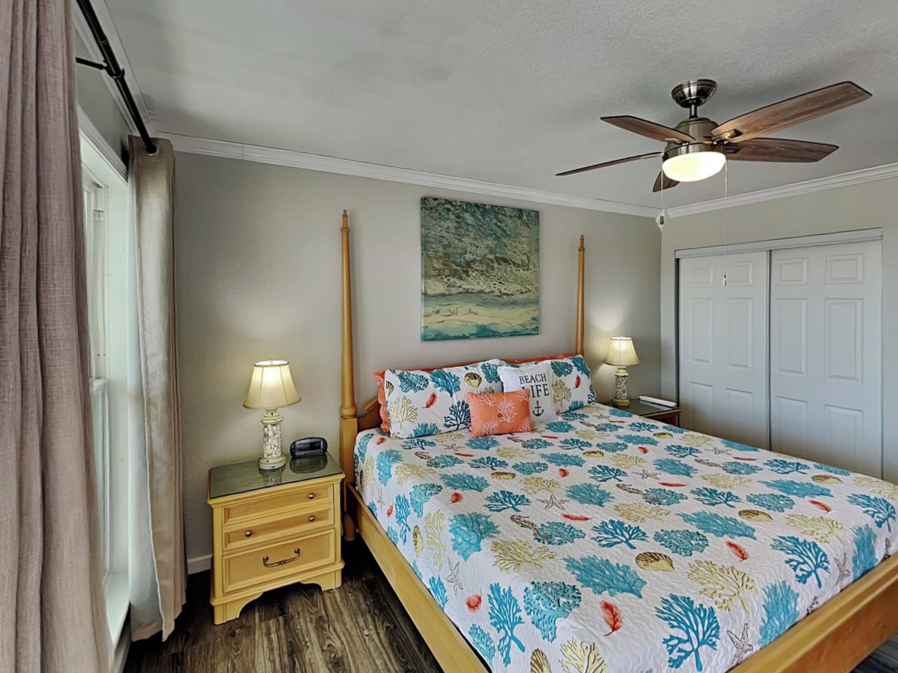 A bedroom with a bed, two bedside tables, a ceiling fan, a painting, and a wardrobe.
