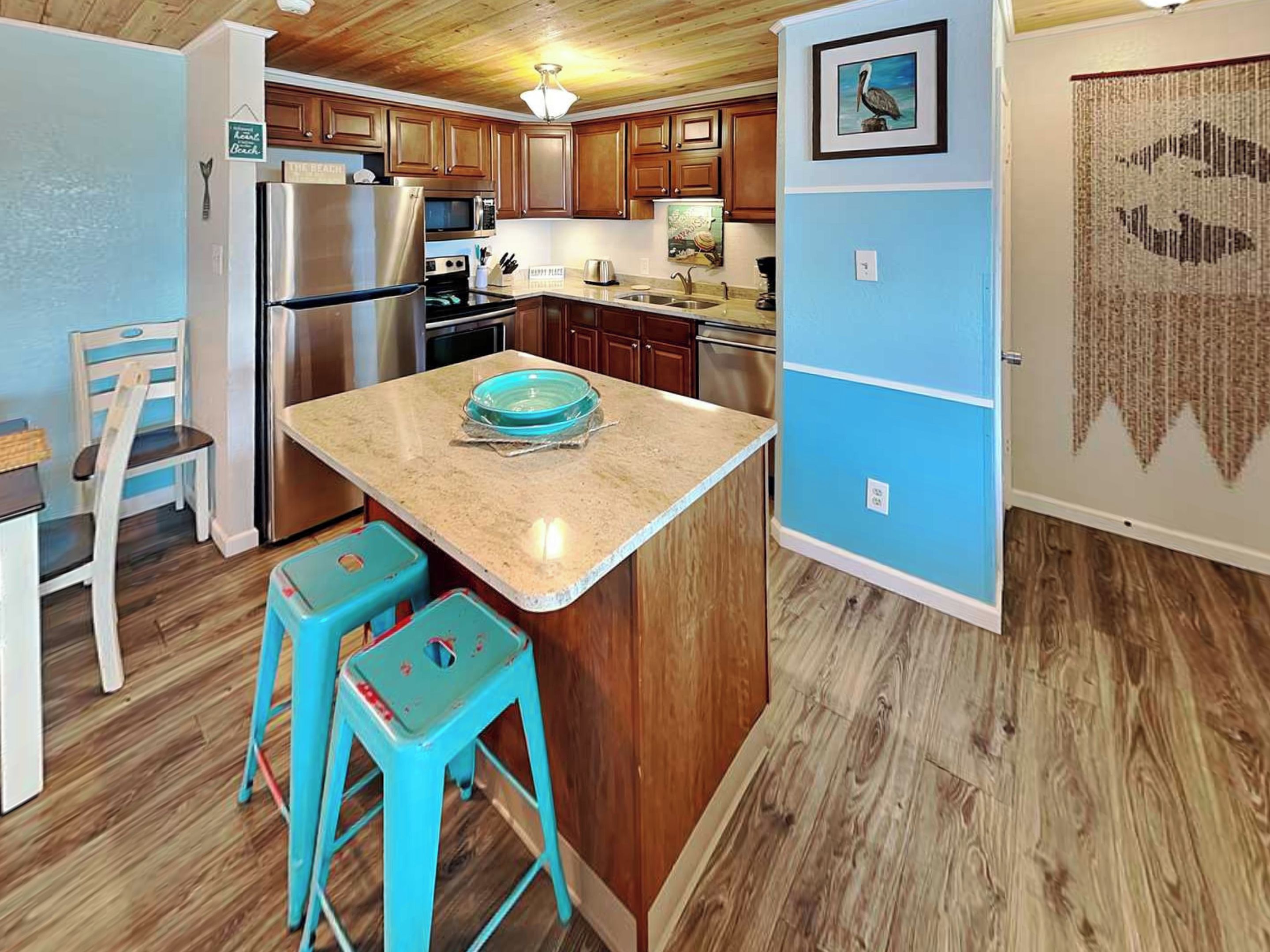 A kitchen with wooden cabinets, a stainless steel refrigerator, a microwave, and a sink. There are two turquoise stools and a blue bowl on the counter.
