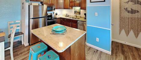 A kitchen with wooden cabinets, a stainless steel refrigerator, a microwave, and a sink. There are two turquoise stools and a blue bowl on the counter.