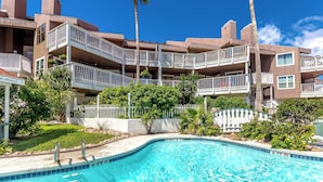 A swimming pool with a 5 FT depth marker, surrounded by a white fence and palm trees, with multi-story buildings in the background. - My Happy Place by Silver Sands Vacation Rentals (Port Aransas)