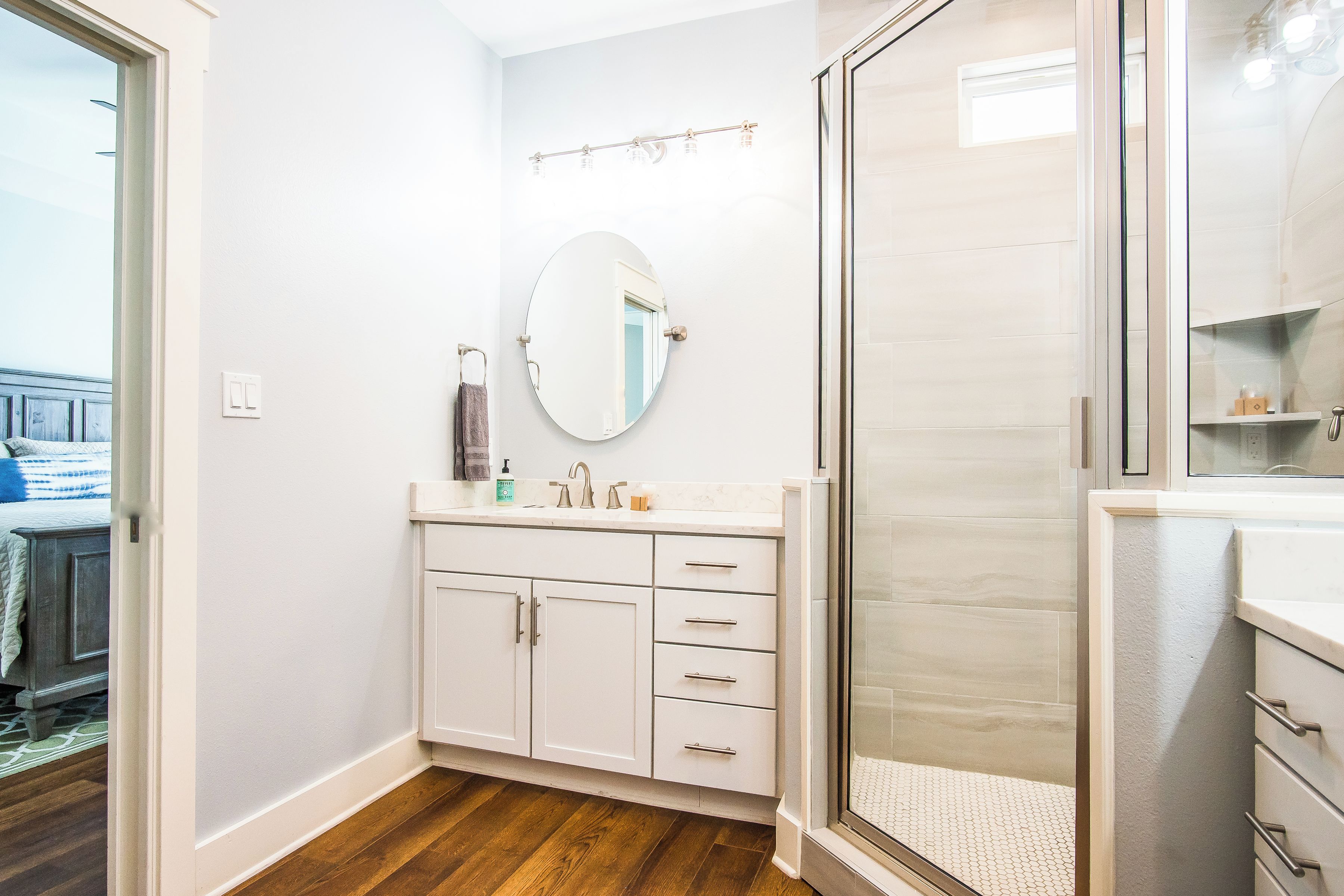 Une salle de bain avec un meuble-lavabo blanc, un miroir rond, une cabine de douche en verre et un plancher en bois.