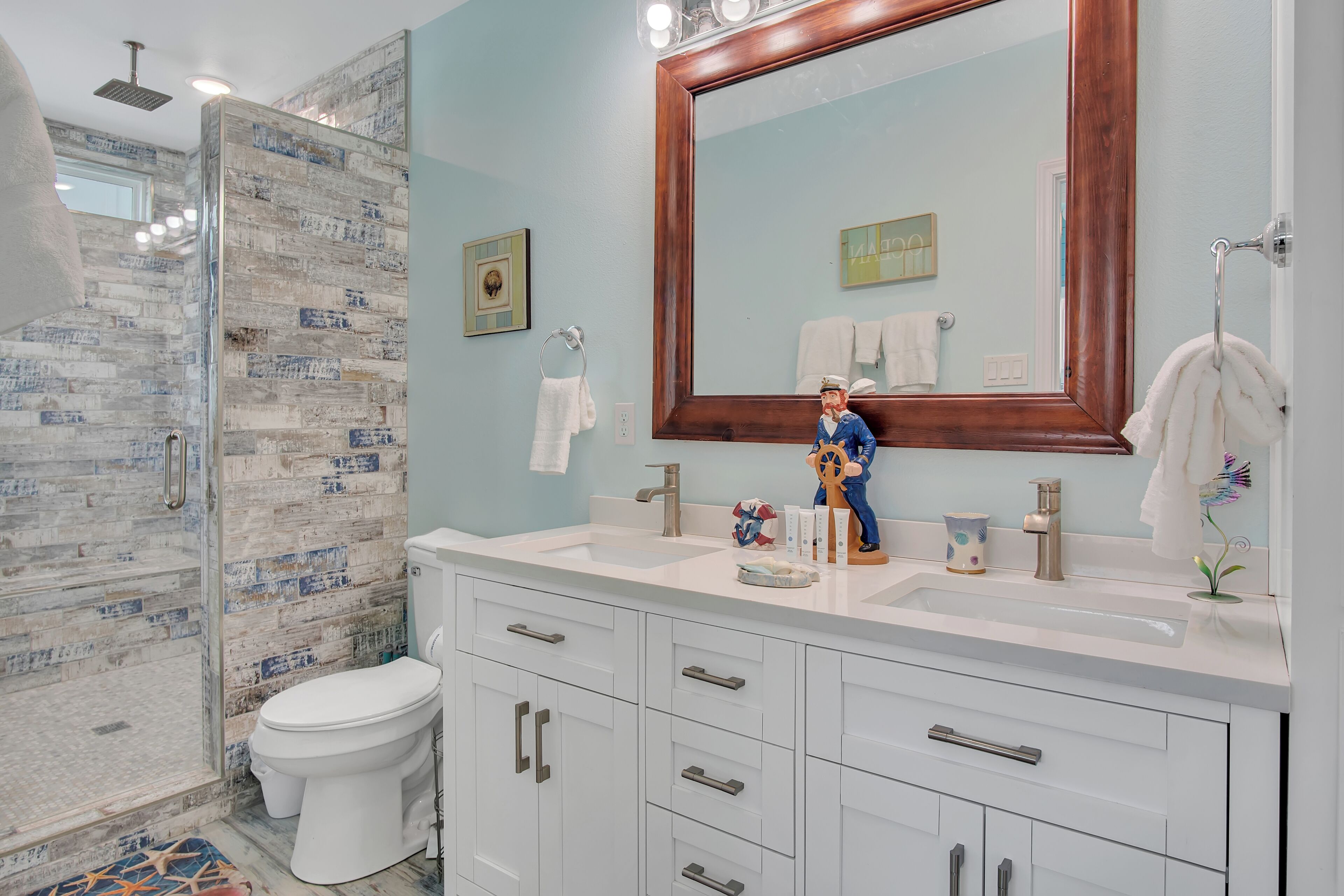 Un baño con cabina de ducha de vidrio, un inodoro blanco, un tocador con un espejo grande y una figura decorativa en el lavabo.
