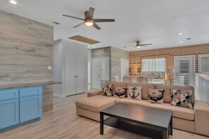 A modern living room with a beige sectional sofa, a black coffee table, and a kitchen area with blue cabinets.