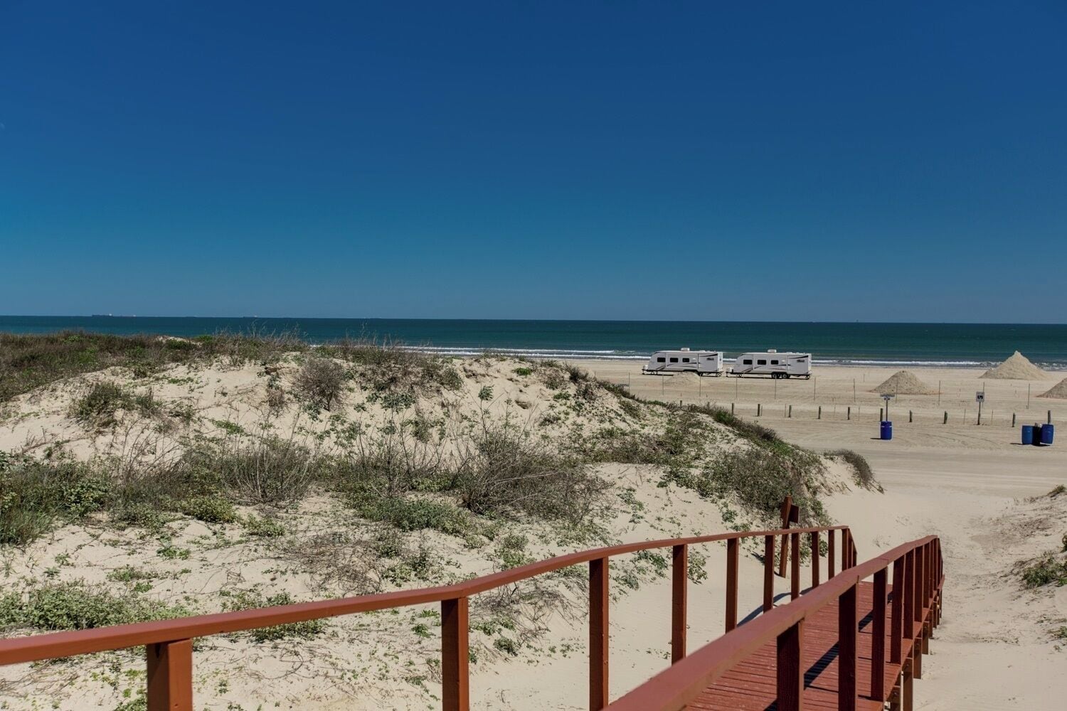 Une plage avec une promenade en bois qui mène à une zone sablonneuse o ù sont garés des véhicules de loisirs.
