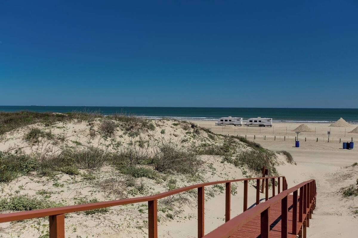 Una playa con un paseo marítimo de madera que conduce a una zona arenosa con vehículos recreativos estacionados.