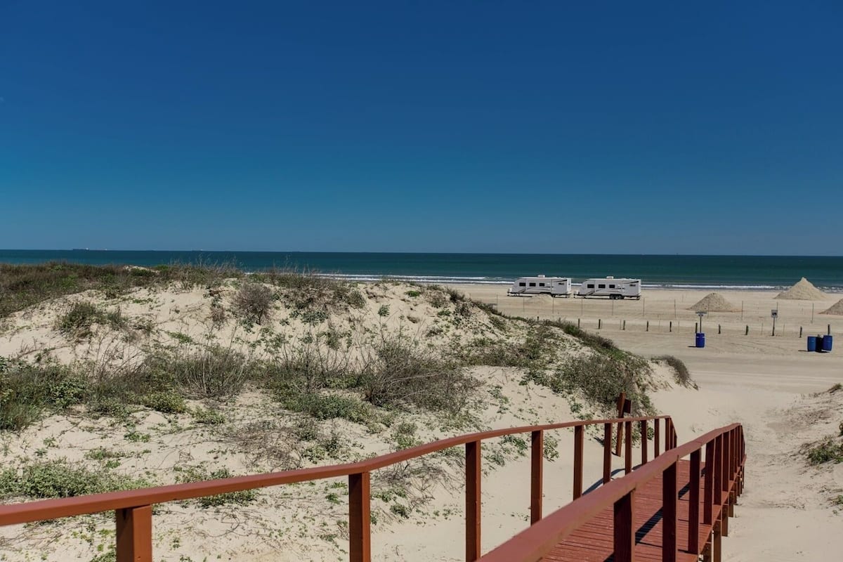 A beach with a wooden boardwalk leading to a sandy area with RVs parked.