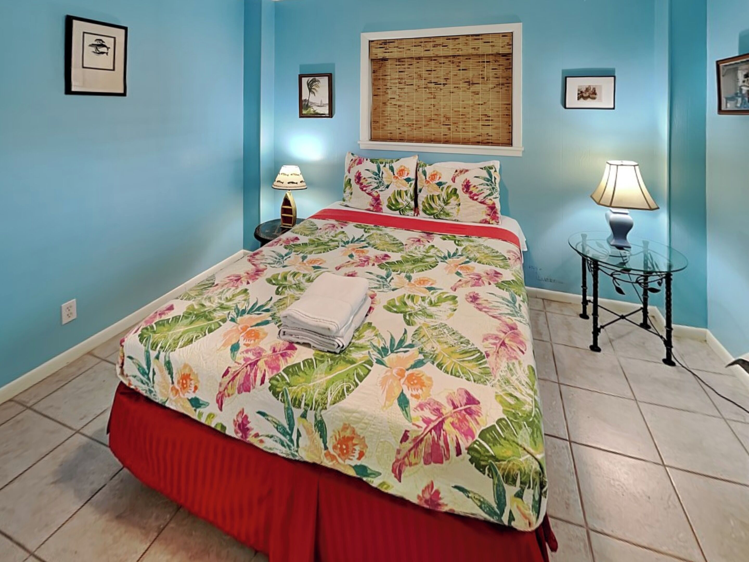A bedroom with a floral bedspread, a red skirted bed, two bedside lamps, a small round table, and a window with a bamboo blind.