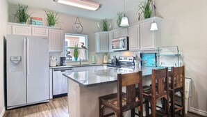 A kitchen with white cabinets, a refrigerator, a microwave, and a sink. There are plants on the cabinets and a clock on the wall. - Port A Good Time by Silver Sands Vacation Rentals (Port Aransas)
