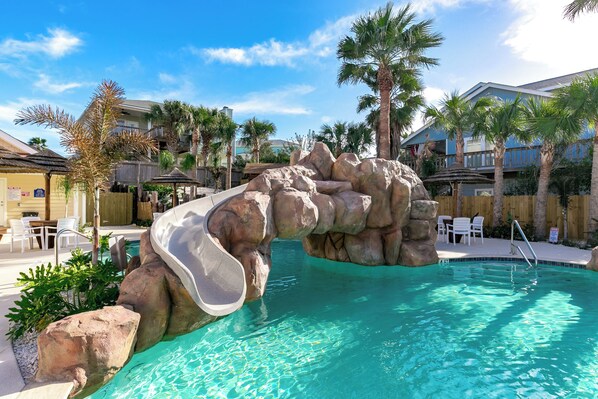 A pool with a rock slide, palm trees, and lounge chairs. - Port A Good Time by Silver Sands Vacation Rentals (Port Aransas)