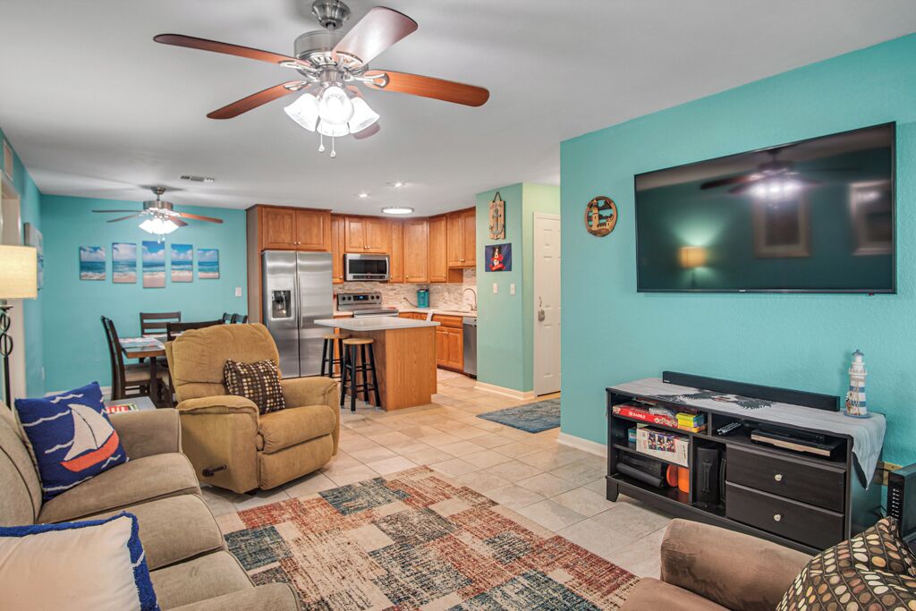 A living room with a sofa, a coffee table, and a TV. There is a kitchen area with a refrigerator, microwave, and stove. The room has a ceiling fan and a rug on the floor.