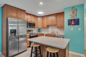 A kitchen with wooden cabinets, stainless steel appliances, a white island with stools, and a blue accent wall. - Pawsitively Paradise by Silver Sands Vacation Rentals (Port Aransas)