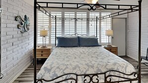 A four-poster bed with a floral bedspread, two bedside tables with lamps, a ceiling fan, and a brick wall with a decorative octopus sculpture. - Pearl Marlin by Silver Sands Vacation Rentals (Port Aransas)