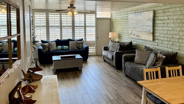 A spacious living room with two sofas, a coffee table, and a wooden dining table. Large windows provide ample natural light, and a ceiling fan is visible. - Pearl Marlin by Silver Sands Vacation Rentals (Port Aransas)