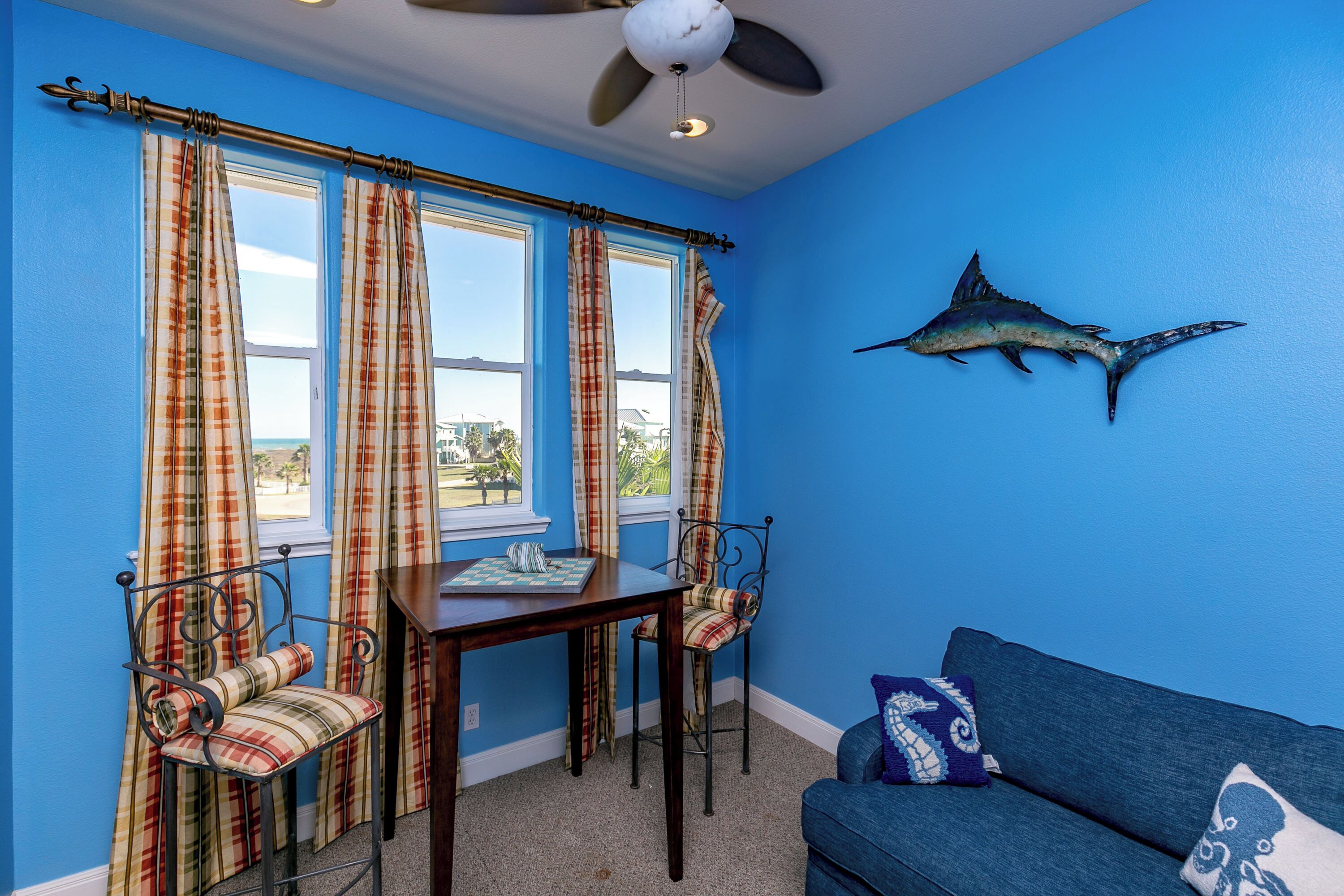 A room with blue walls, a ceiling fan, plaid curtains, a dining table with chairs, and a blue sofa.