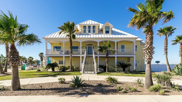 A two-story yellow building with a white balcony, palm trees, and a clear blue sky. - Porpoise of Life by Silver Sands Vacation Rentals (Port Aransas)