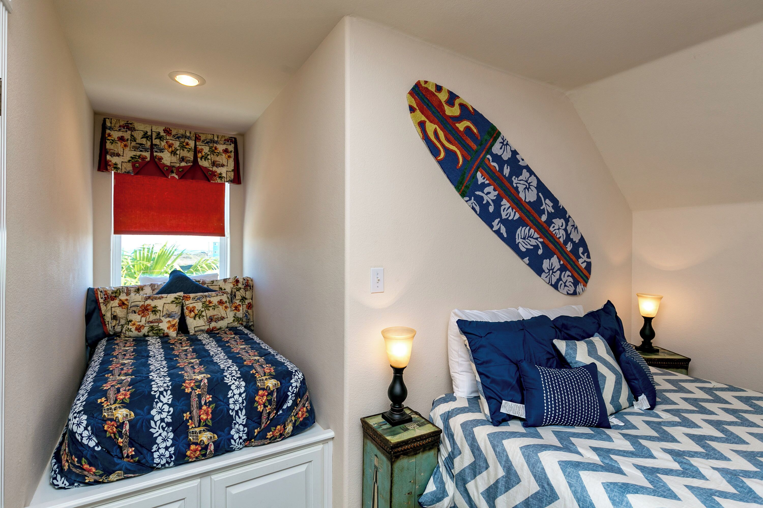 Two beds with patterned bedding, a window with a red blind, and a wall-mounted decorative piece.