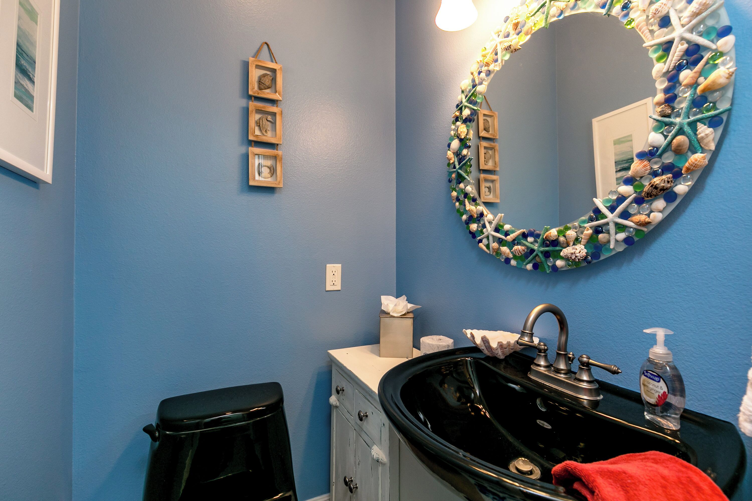 Un baño con pared azul, lavamanos negro, espejo con detalles de concha marina y estrella de mar, y cuadros en la pared.