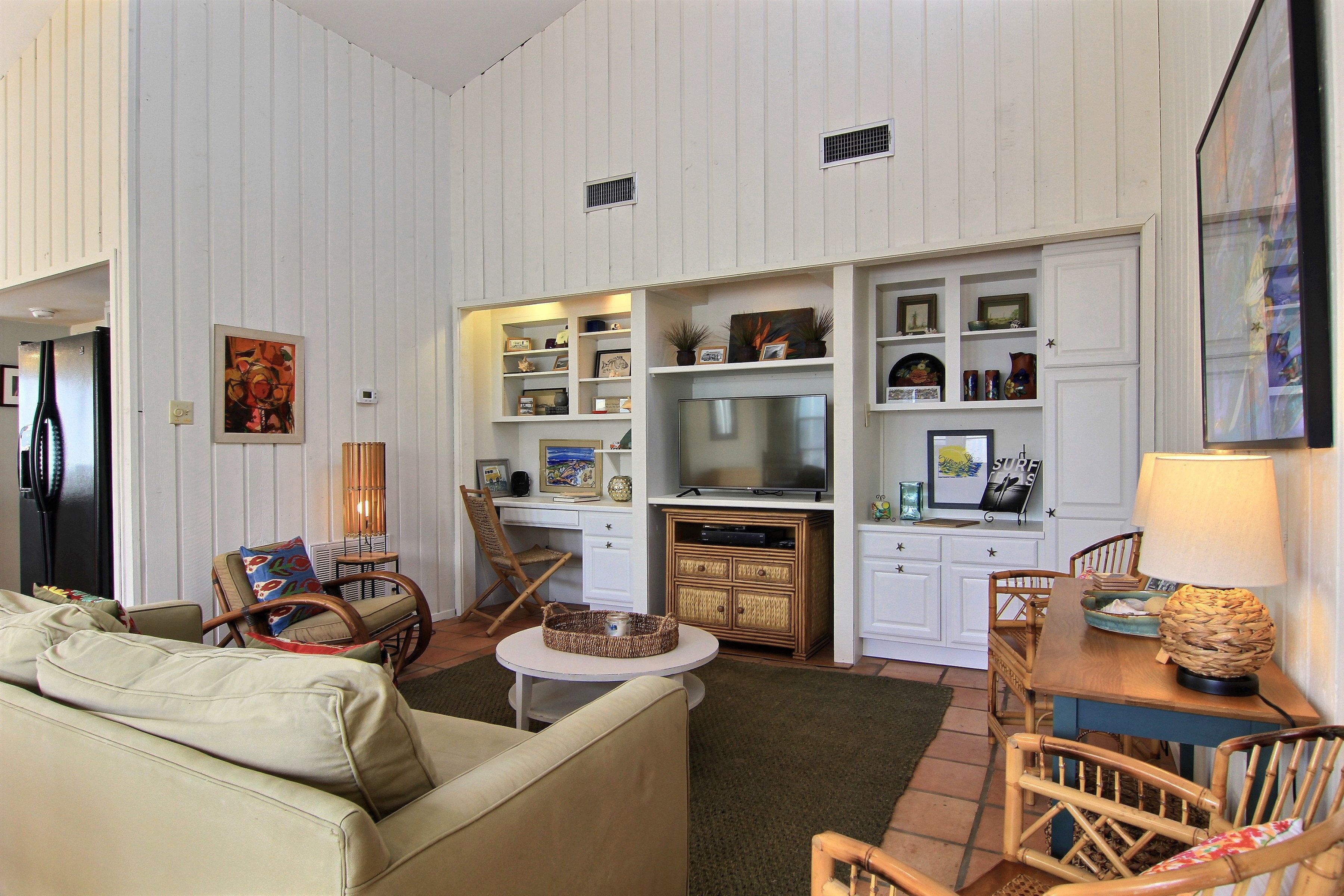 A living room with a sofa, chairs, a TV, a dining table, and a kitchen area with cabinets and a refrigerator.