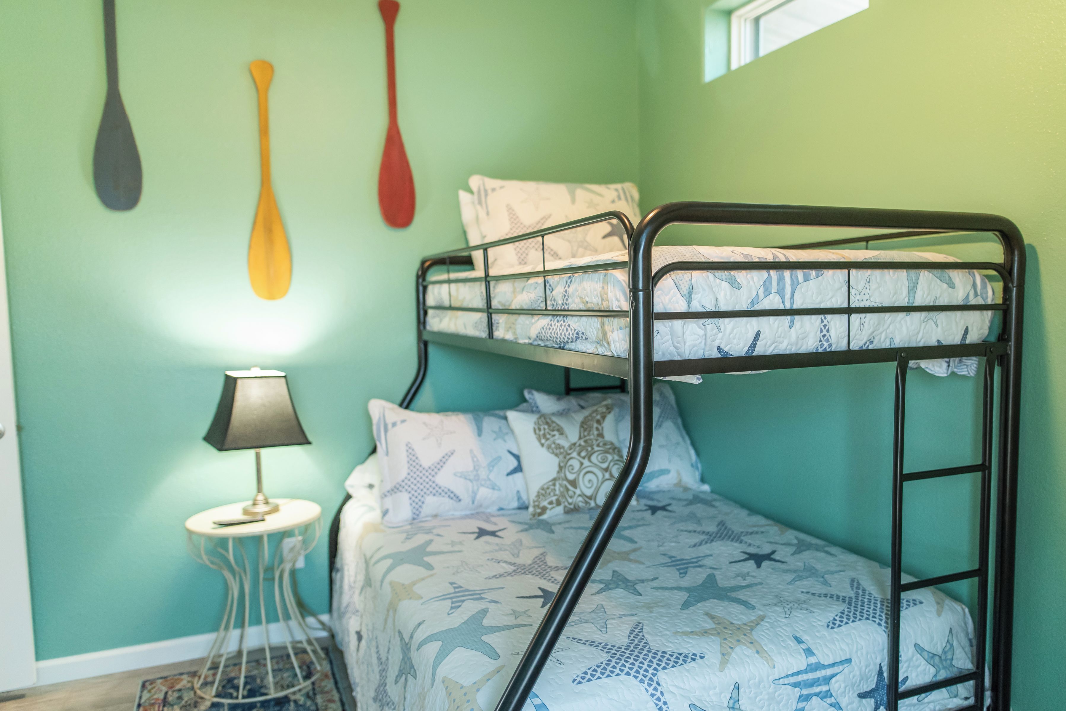 Un lit superposé avec une couverture à motifs d’étoiles, une petite table de chevet avec une lampe et deux pagaies fixées au mur.