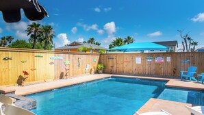 A swimming pool with a wooden fence decorated with fish cutouts, a blue umbrella, and a sign warning against lifeguard presence. - Port A Ponderosa by Silver Sands Vacation Rentals (Port Aransas)