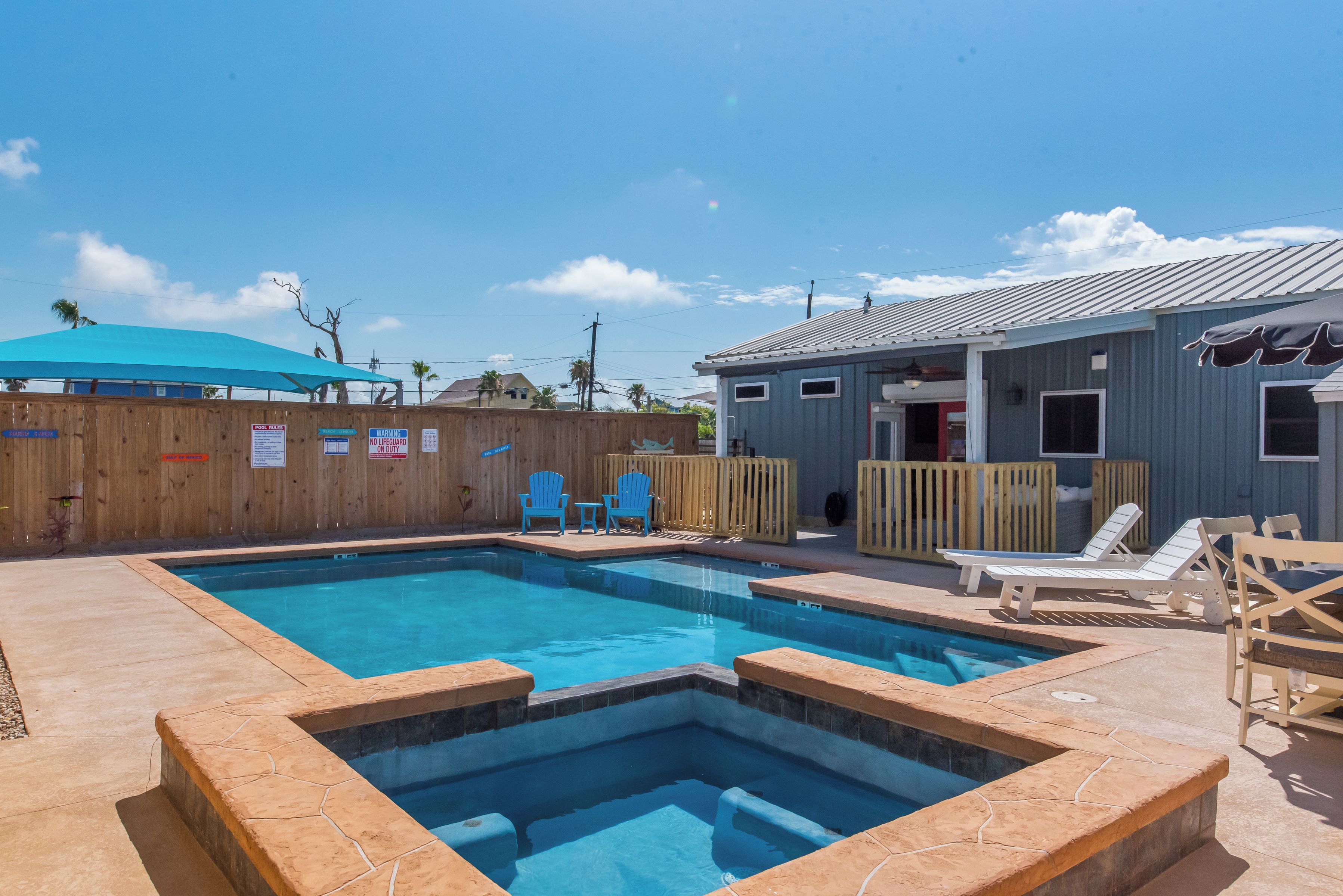 A swimming pool with a wooden deck, lounge chairs, and a covered area.