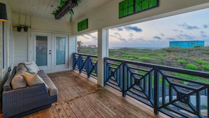 A covered porch with a wooden floor, a cushioned bench, and a view of the beach.