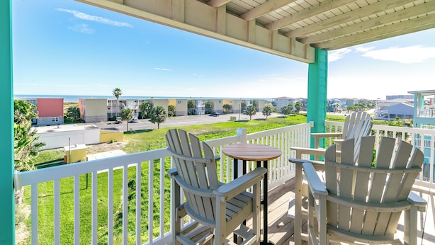 A balcony with two Adirondack chairs and a small table, overlooking a parking lot and buildings. - Port A Paradise by Silver Sands Vacation Rentals (Port Aransas)