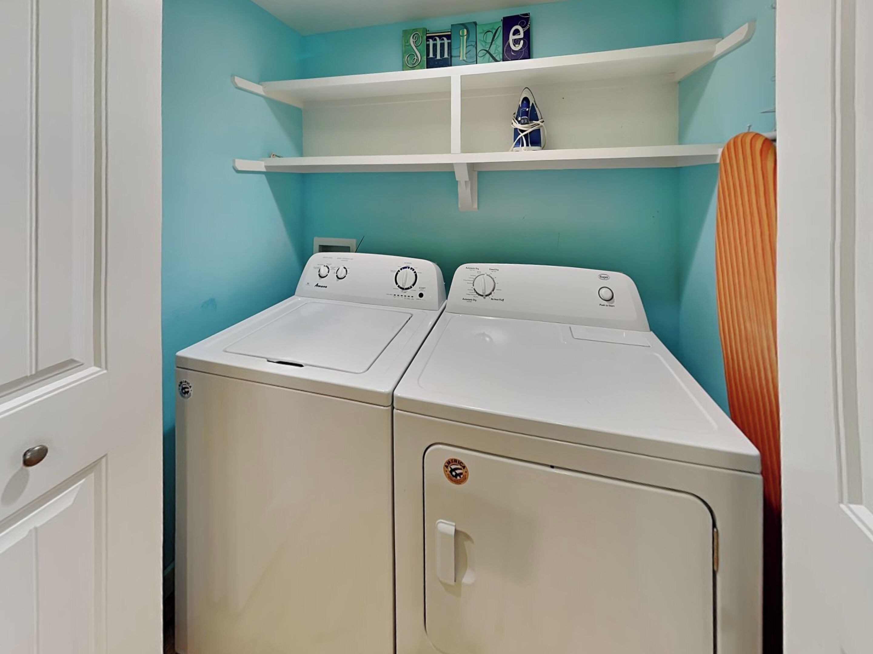 A laundry room with two front-loading washing machines and a dryer. There are white shelves above the machines, and a towel rack to the right.