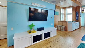 A living room with a TV mounted on a blue wall, a white entertainment unit, wooden flooring, and a kitchen area visible in the background. - Port A Paradise by Silver Sands Vacation Rentals (Port Aransas)