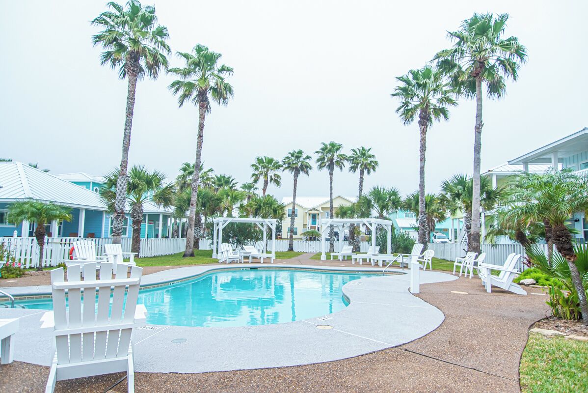 A pool area with palm trees, white chairs, and a covered seating area.