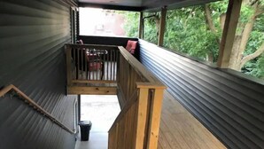 Eine Holztreppe führt zu einem Balkon mit Metallverkleidung und Blick auf grüne Vegetation.