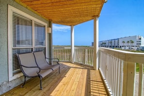 A wooden deck with a bench, white railings, and a view of the beach and buildings in the background.