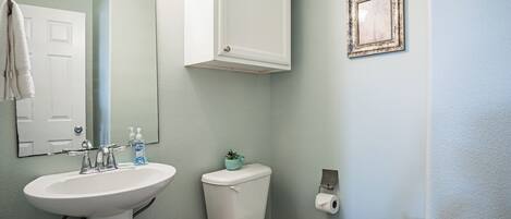 A bathroom with a white sink, toilet, and a mirror. There is a framed picture on the wall and a small plant on the toilet tank.