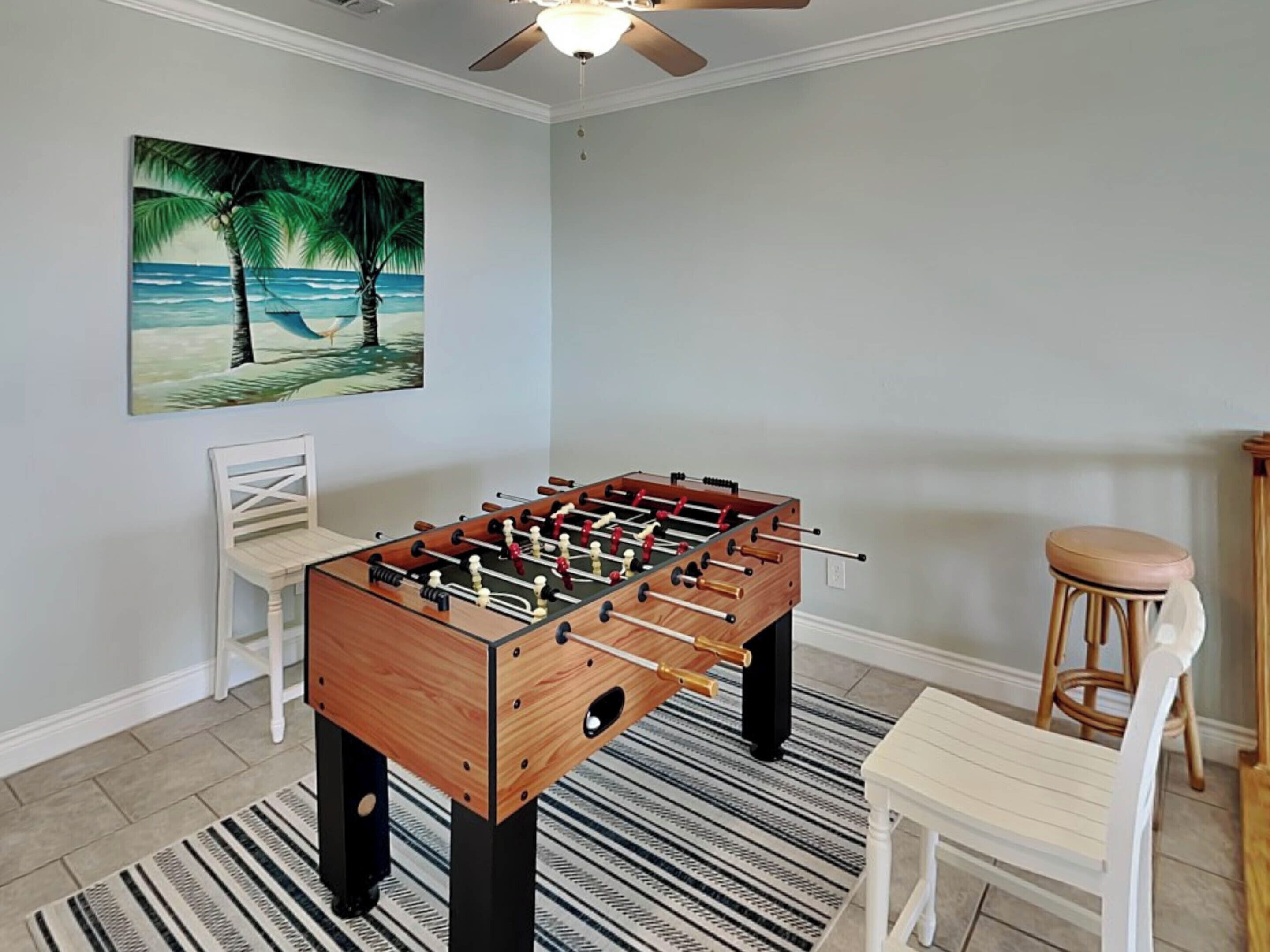 Ein Zimmer mit einem Kicker, einem Stuhl, einem Hocker, einem Strandbild und einem Deckenventilator.