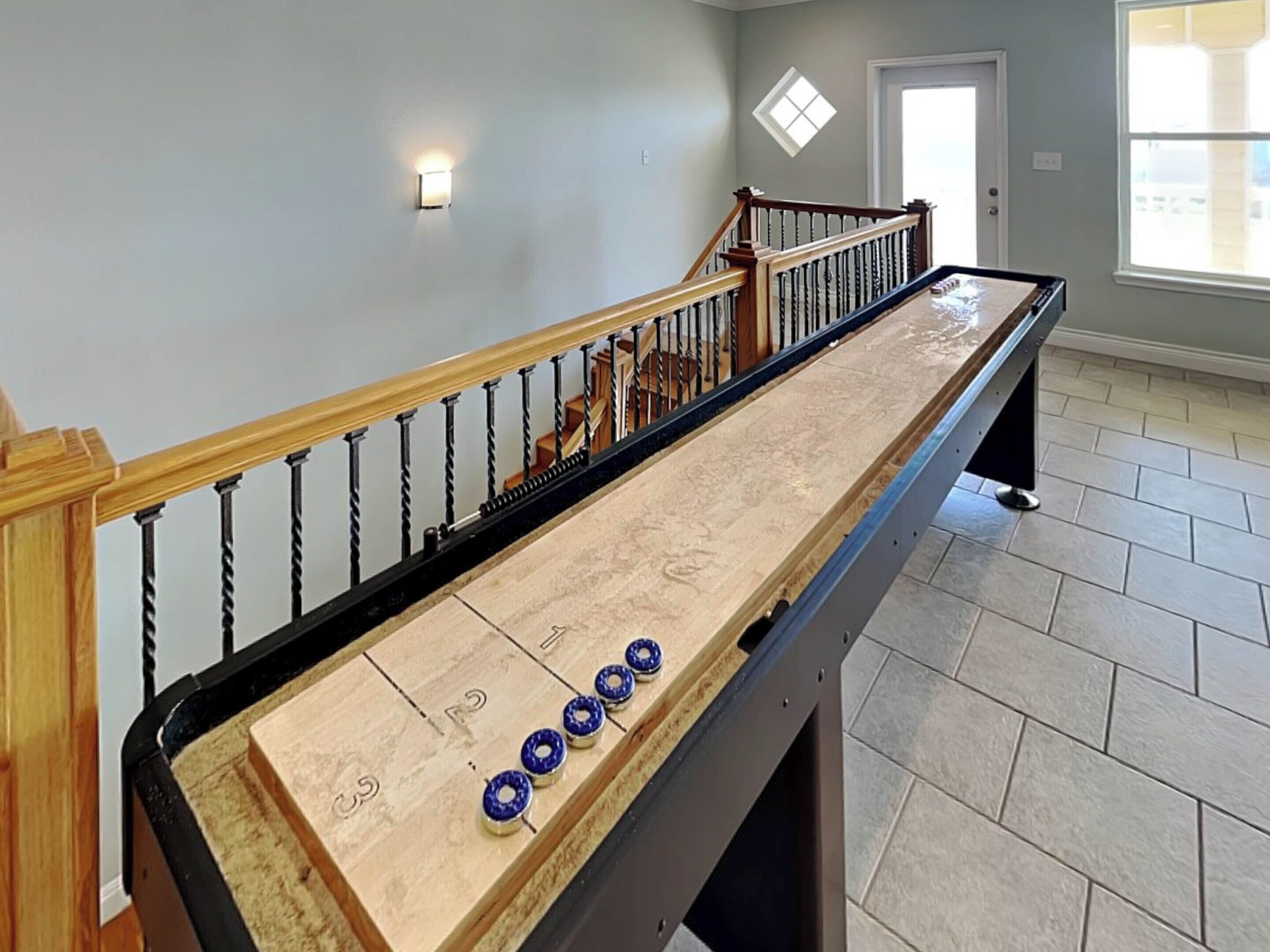 A shuffleboard table in a room with a wooden staircase and a small window.