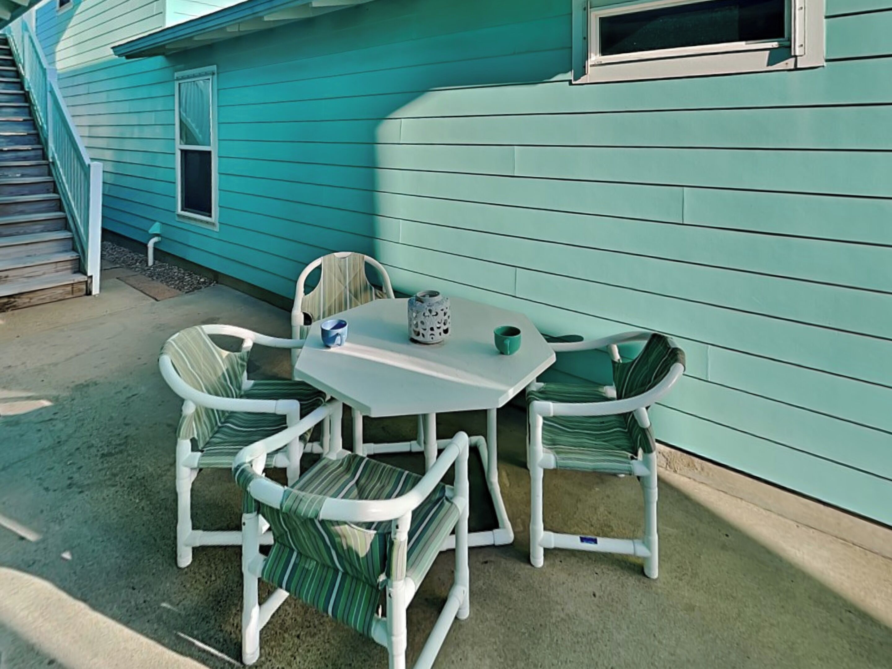 An outdoor patio with a white table and four chairs, a small table lamp, and a window with a curtain.