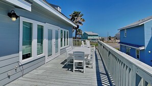Une terrasse en bois avec une table et des chaises, donnant sur une plage où l’on aperçoit des maisons au loin.