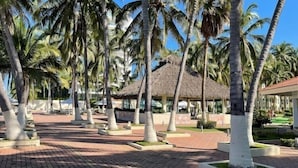 A resort area with palm trees, a thatched-roof structure, and a paved walkway. - Costa Vallarta by TRVL2HM (Nuevo Nayarit)