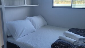 Een compact bed met witte lakens en een zwart hoofdbord, een raam met uitzicht op heuvels, en een gebloemd deken.
