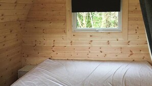 Une petite pièce en bois, aménagée comme une cabane, avec un lit simple, une fenêtre et une vue sur les arbres à l’extérieur.