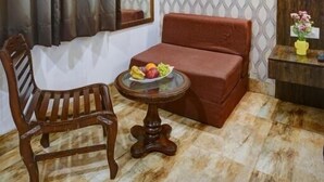 A room with a wooden chair, a small round table with fruit, a cushioned bench, a mirror, patterned wallpaper, and a wall-mounted light fixture. - Hotel Nirvana-Near IGI Airport Delhi (New Delhi)