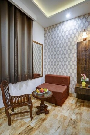 A room with a wooden chair, a small round table with fruit, a cushioned bench, a mirror, patterned wallpaper, and a wall-mounted light fixture. - Hotel Nirvana-Near IGI Airport Delhi (New Delhi)