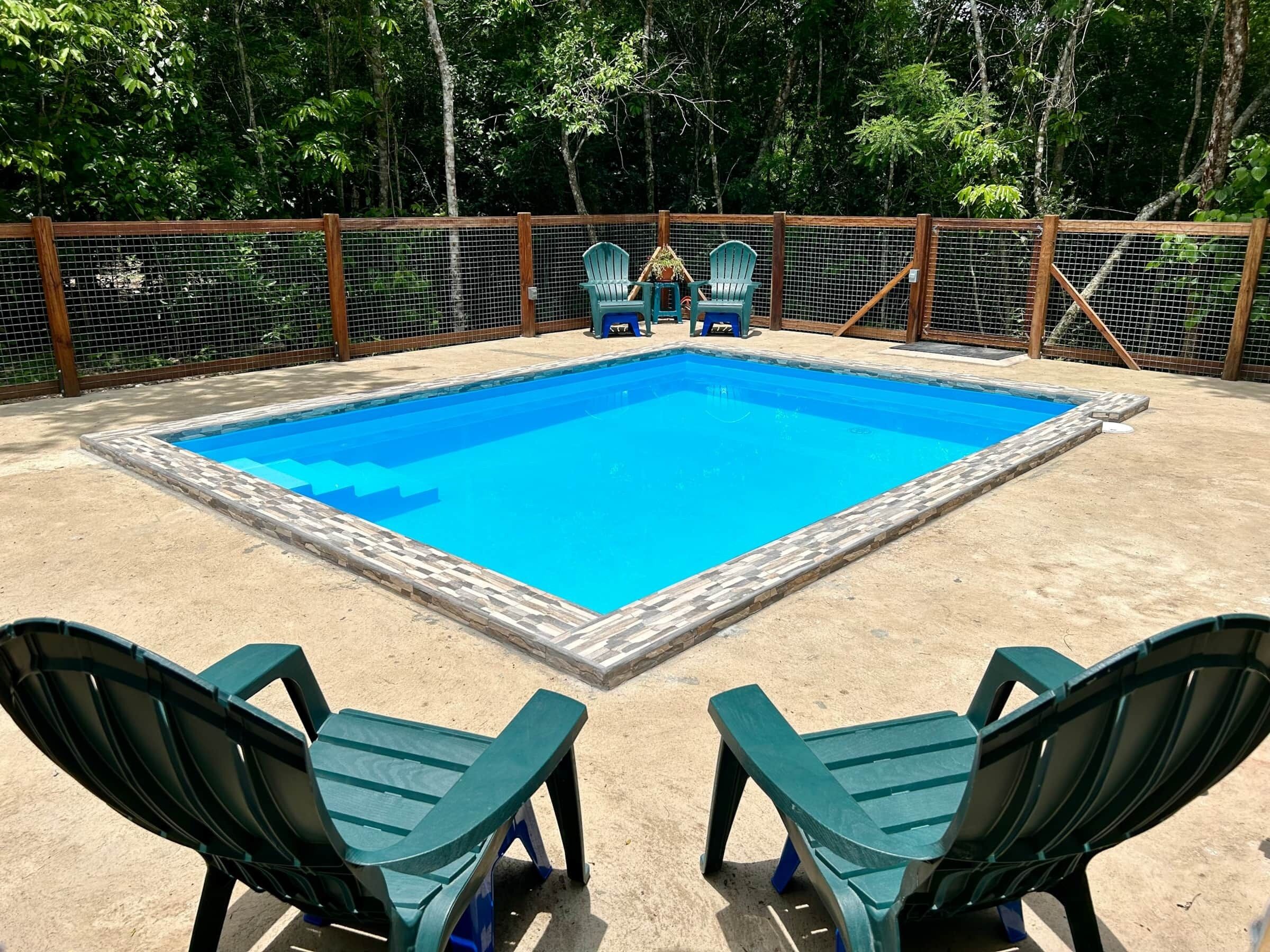 A rectangular swimming pool with a tiled border, surrounded by a wooden fence and a sandy deck.