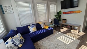 A living room with a blue sofa, patterned cushions, a TV mounted on the wall, and a surfboard decoration.