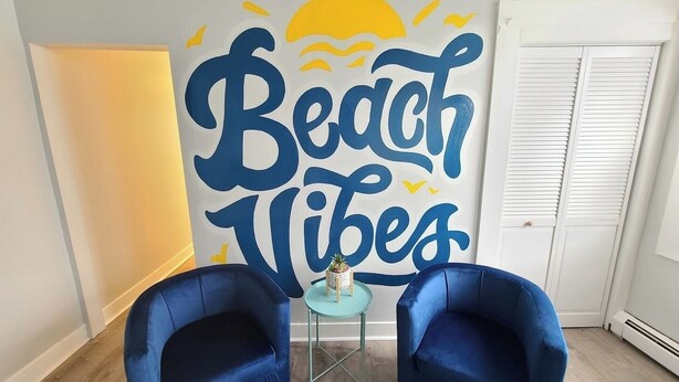 A room with a large wall mural saying "Beach Vibes," two blue armchairs, a small round table with a plant, and a white door with shutters.